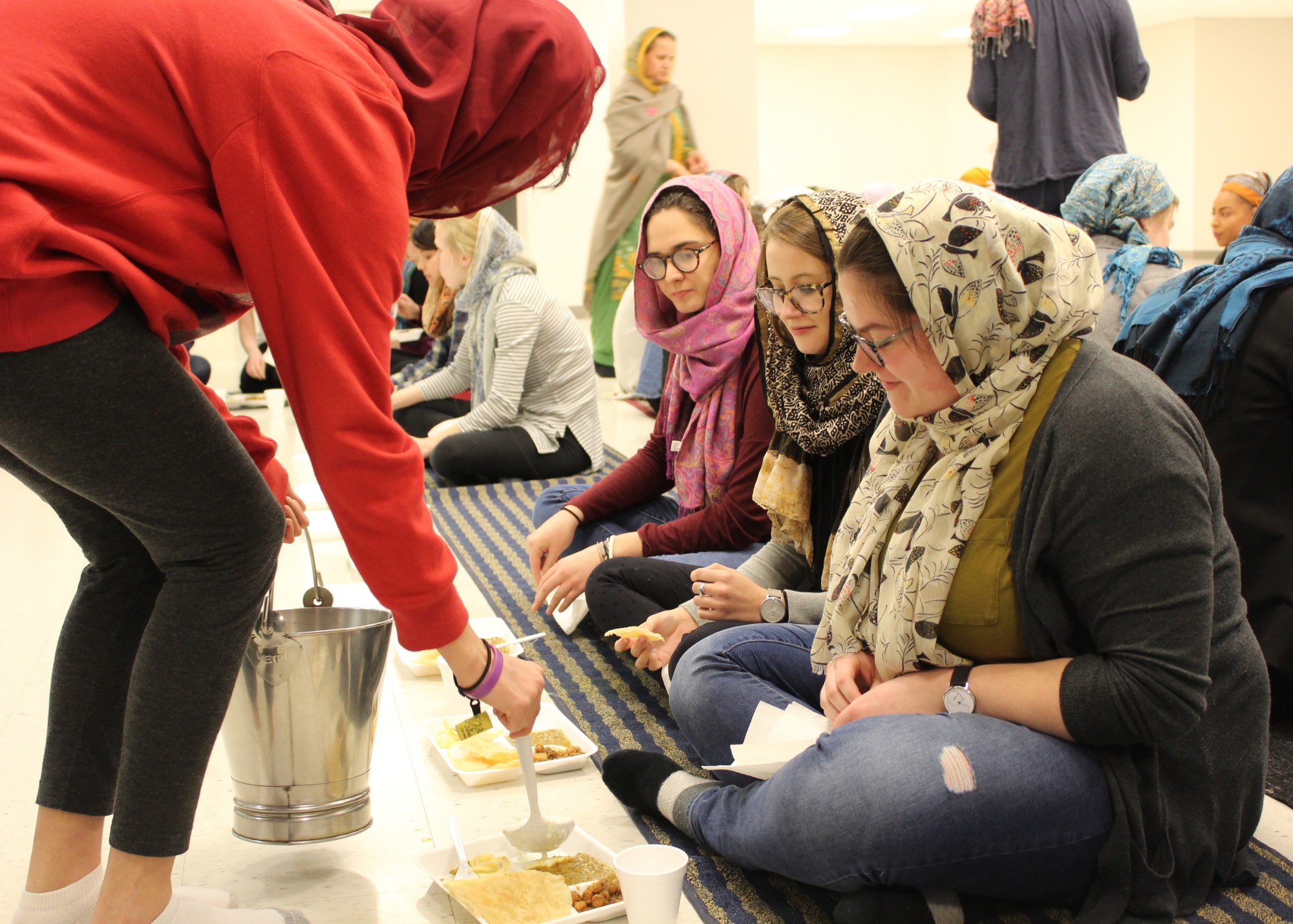 Students with headscarves sitting on the floor of a gurdwara or Sikh temple being fed langar (the shared communal meal) by a volunteer
